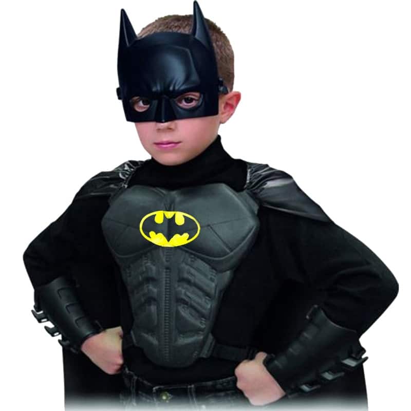 Enfant qui porte un déguisement de batman composé du masque du héro, d'une plaque sur la poitrine, de protections de poignets, et d'une cape , il a les mains posées sur les hanches