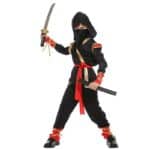 Enfant portant le costume de Ninja pour enfant, il est noir et rouge et comprend une tunique, un pantalon, une capuche, une ceinture, un masque et 4 rubans dorés, sur fond blanc