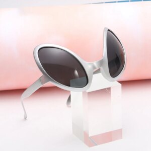 Lunettes en forme des yeux d'extraterrestre, aux verres noires et monture argentée. Les lunettes sont sur une plaque en acrylique sur un fond rose.