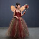 Une petite fille porte un déguisement de fée. Ce déguisement est composé d'une robe en tulle avec des couleurs de la forêt. Le costume est composé également d'une baguette, d'ailes orange et d'oreilles de fée.