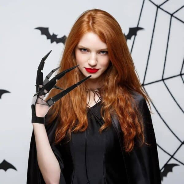 Femme déguisée pour Halloween habillée en noir avec un accessoire de main avec des doigts articulés noirs, sur fond gris